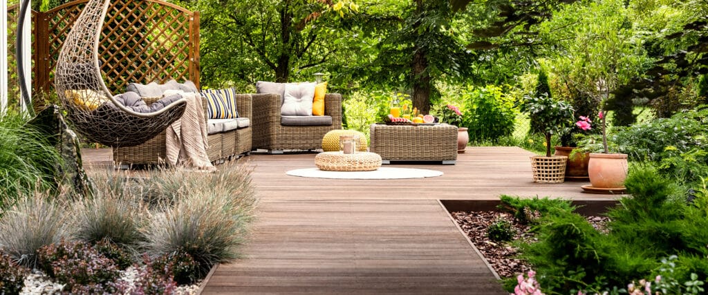 Terrasse bois dans jardin luxuriant, chaise suspendue et salon extérieur avec fruits/boissons sur table.