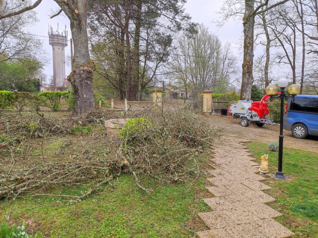 Jardin avec tas de branches fraîchement coupées et un broyeur rouge près d'un van bleu. Tour d'eau et chemin pavé.